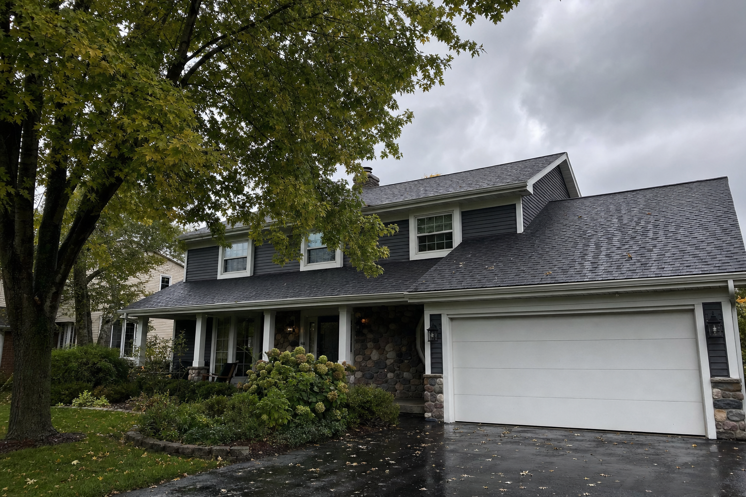 documentary photo from homeowner POV at ground level looking up at a Lake County asphalt shingle roof after a storm, damp driveway, mature maple tree, gutters and fascia visible, overcast Lake Michigan moody gray sky, no people, no text on image