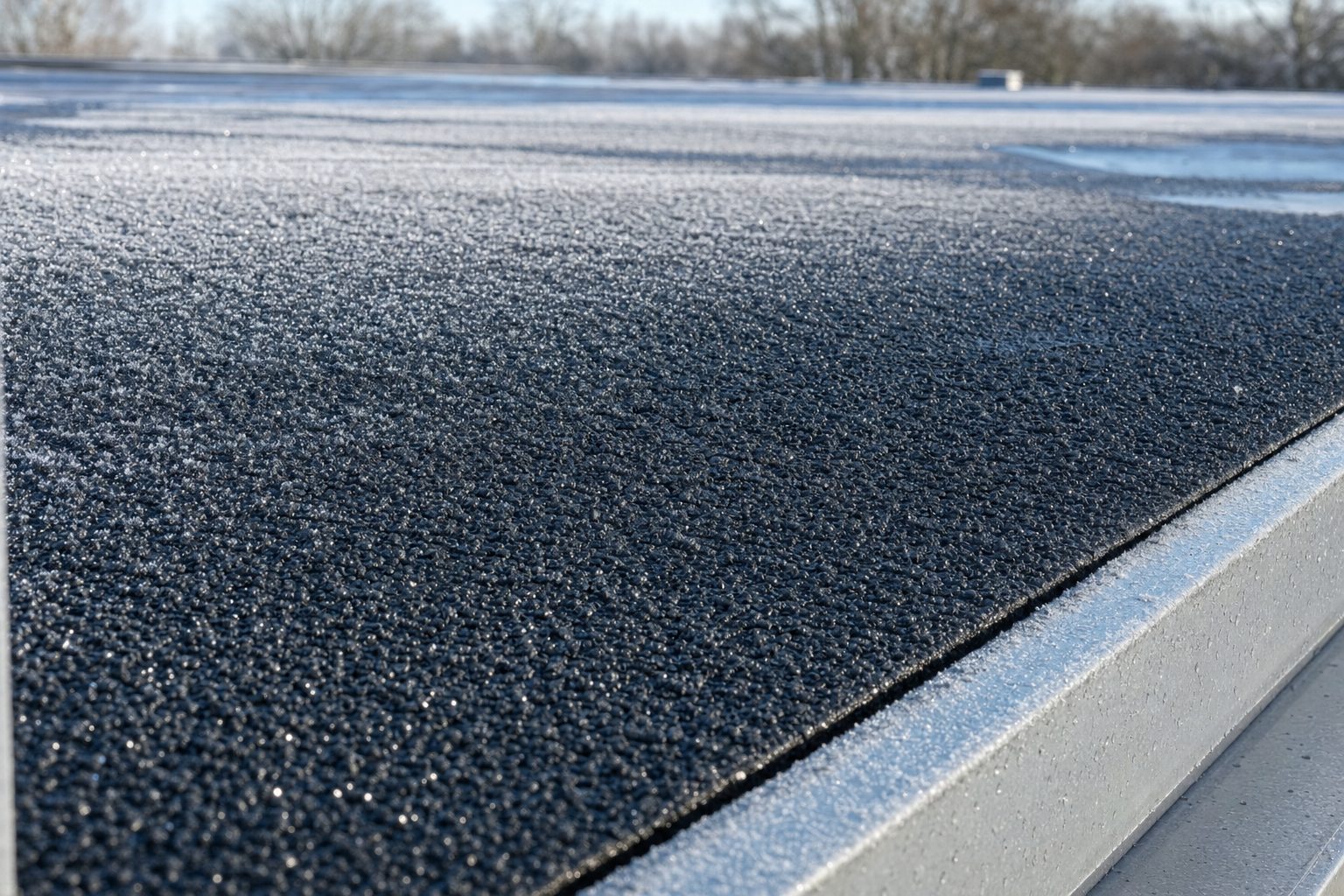 primer plano realista de una membrana granular de betún modificado en un techo de baja pendiente, escarcha ligera en la superficie, borde metálico visible, luz fría de invierno, sin personas, sin texto visible